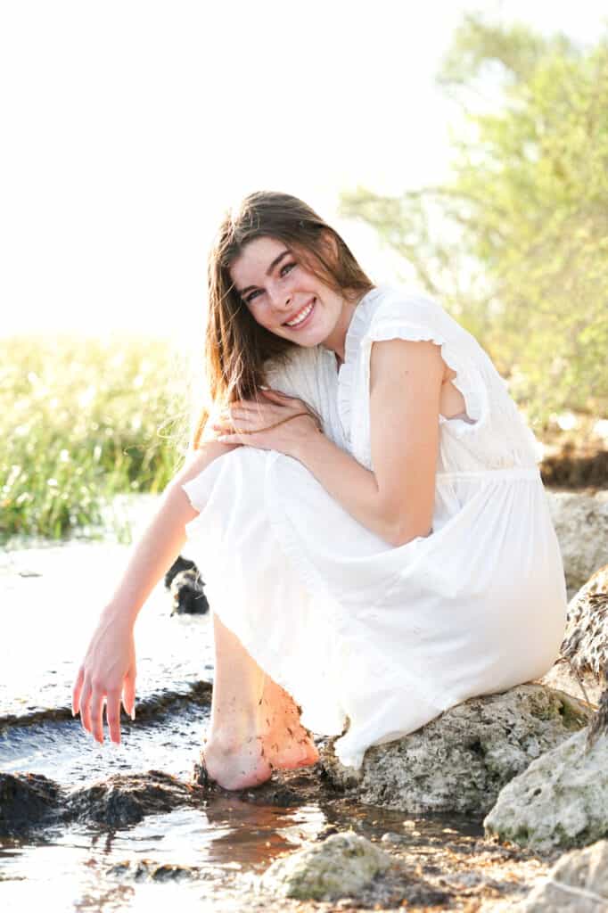 A girl in a white dress is sitting on rocks in the water.