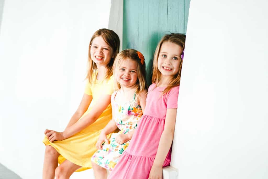 Three little girls in colorful dresses sitting on a window sill.